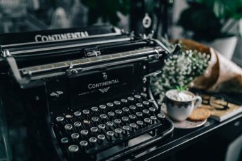 Close-up photo of a typewriter with a coffee and flowers in the background. Photo represents the work of a writer making a new Medium profile page.