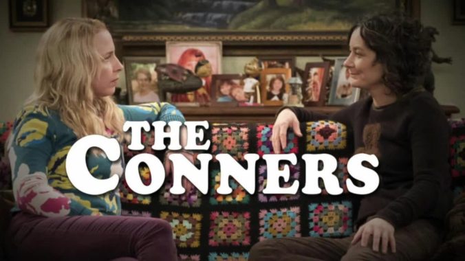 The title of the TV show The Connors, with characters Becky and Darlene sitting on a couch facing one another.