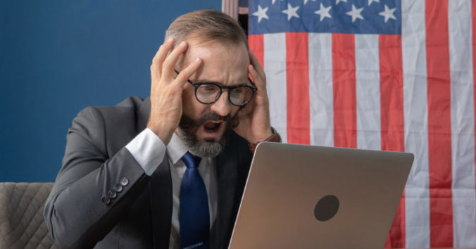 A man sitting in a suit in front of a laptop with a distraught look on his face. A flag sits in the background.