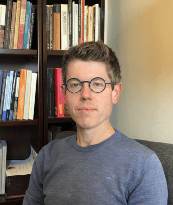 A photo of Matt L. Drabek, a philosophical counselor who is a white man about 40 years old. He is wearing stylish round glasses and a sweater, and there are philosophical books in the background.