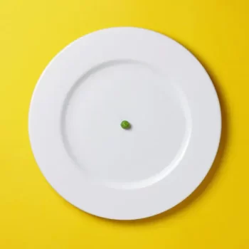 A photo of a white plate against a yellow background with a single green pea in the middle, intended to represent left minimalism.