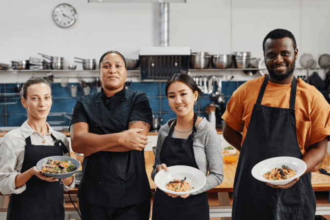 A photo of four restaurant workers holding dishes and smiling. From left to right, they are a white woman, a black woman, an Asian woman, and a black man. They're all wearing restaurant aprons.