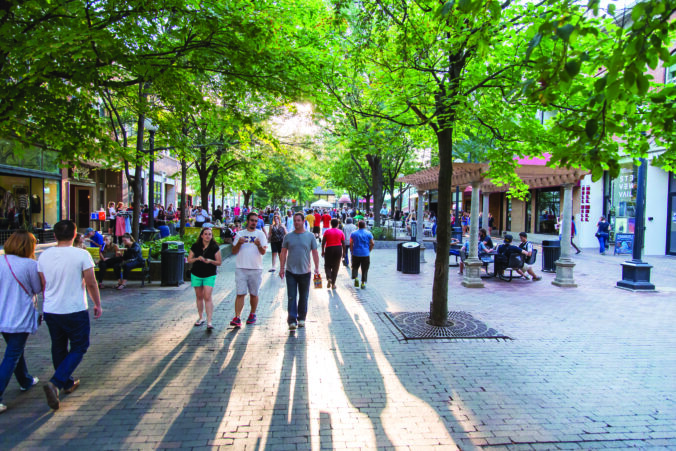 A photo of the Pedestrian Mall (Ped Mall) in Iowa City, Iowa, showing a large pedestrian space with many people walking around on a sunny day with bright green tree leaves.