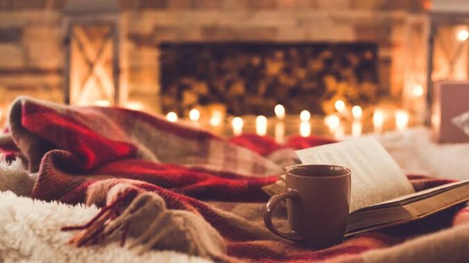 A photo of a cozy room with a coffee mug, book, and blankets in the foreground and candles and a fireplace in the background.