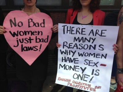 This is a photo of sex worker activists. One holds a heart shaped pink sign reading, "No Bad women just bad laws!" and the other holds a white sign reading, "There are many reasons why women have sex - money is one!"